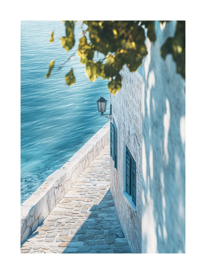 Sunlit narrow walkway by a white stone house overlooking calm ocean water. Ideal for wall art in your home, office, or studio.